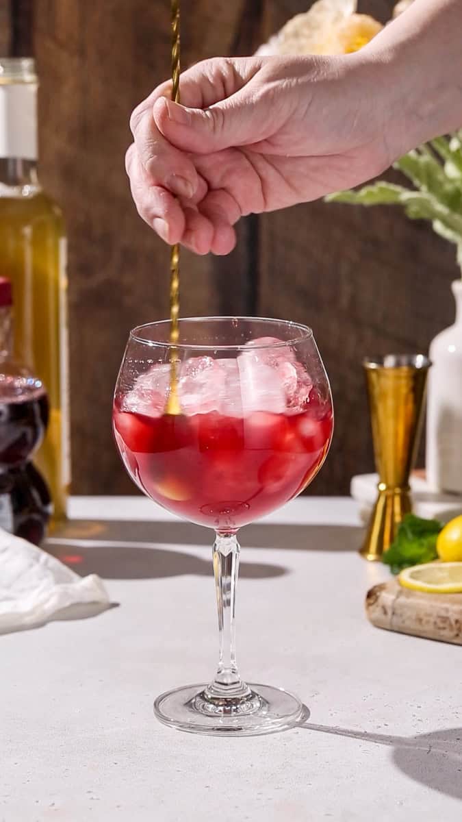 Hand stirring a glass filed with ice and dark red liquid.