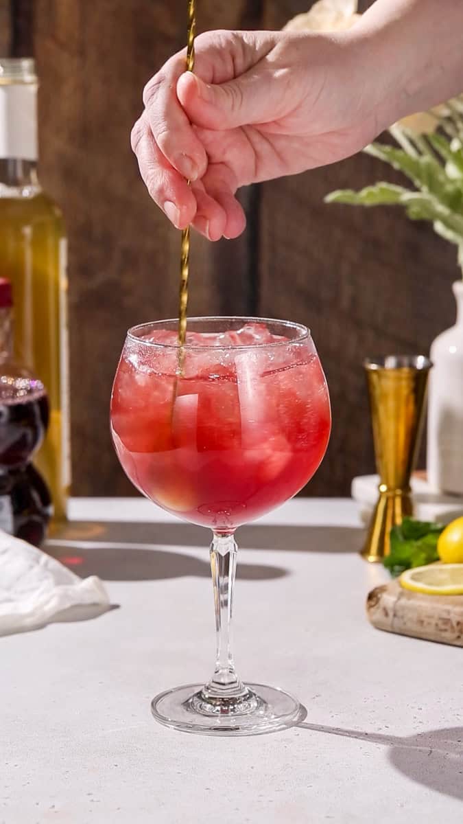 Hand stirring red liquid in a large wine glass with ice.