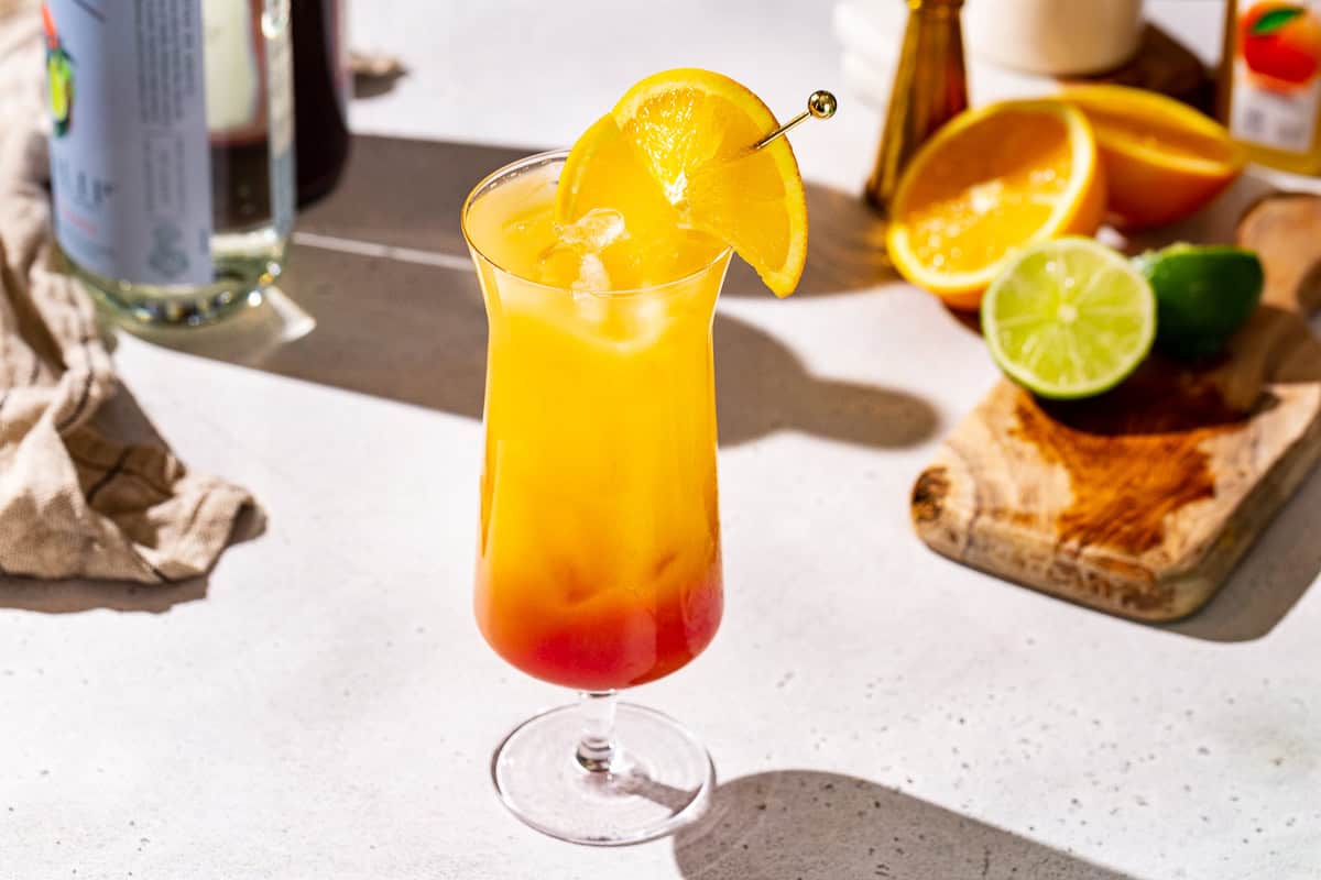 Tequila Sunrise Mocktail on a countertop, garnished with an orange slice. Some bottles and cut orange and lime are in the background.