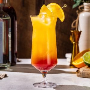 Tequila Sunrise Mocktail with an orange slice garnish on a countertop. The bottom of the drink is red and the top is orange in color.