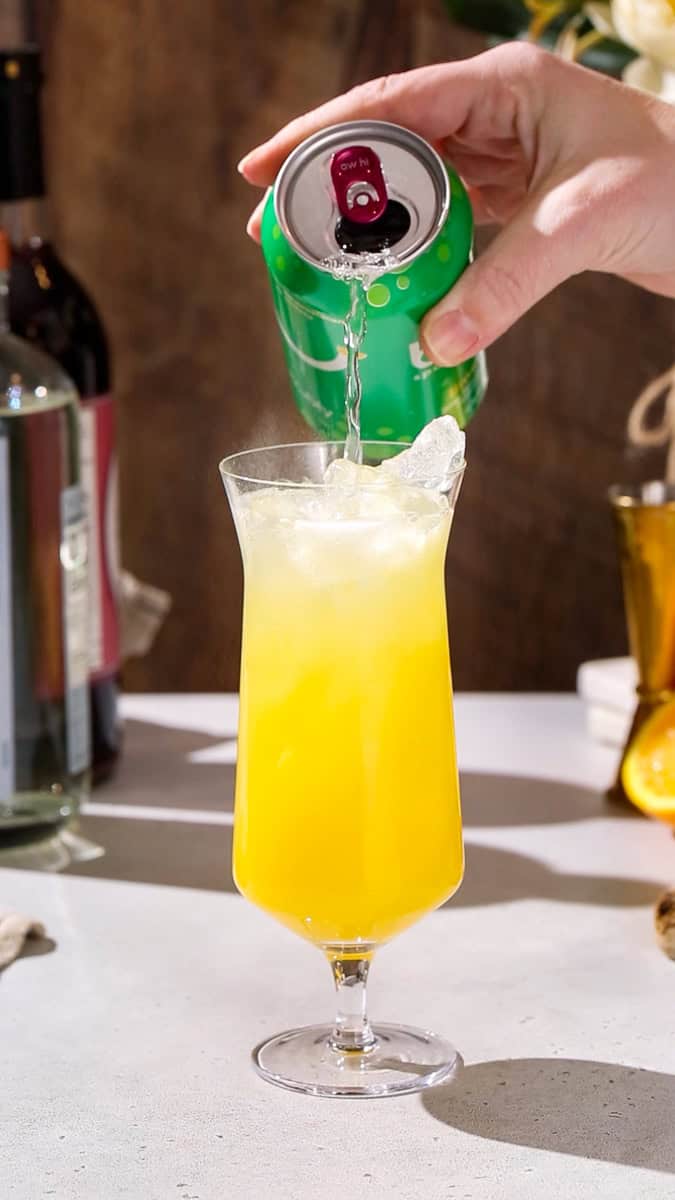 Hand pouring lime seltzer into a cocktail glass filled with orange liquid.