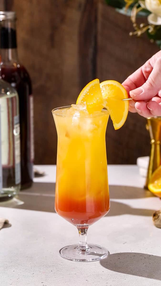 Hand adding an orange slice as garnish to a layered orange and red cocktail.