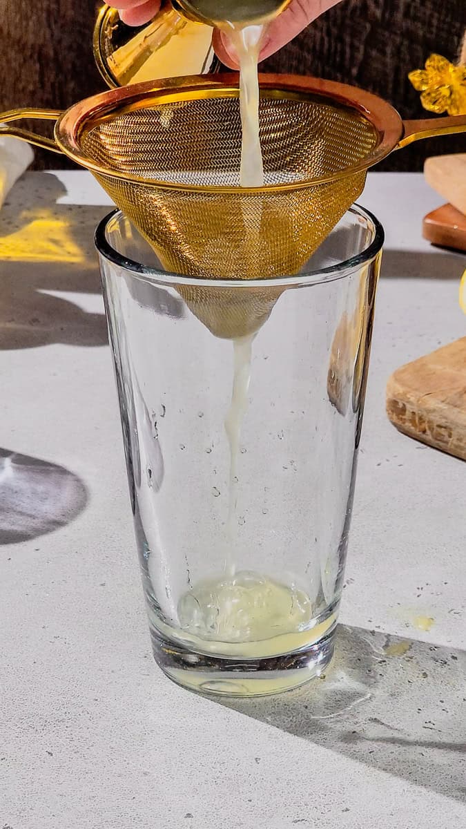 Hand adding lemon juice through a fine mesh strainer into a cocktail shaker.