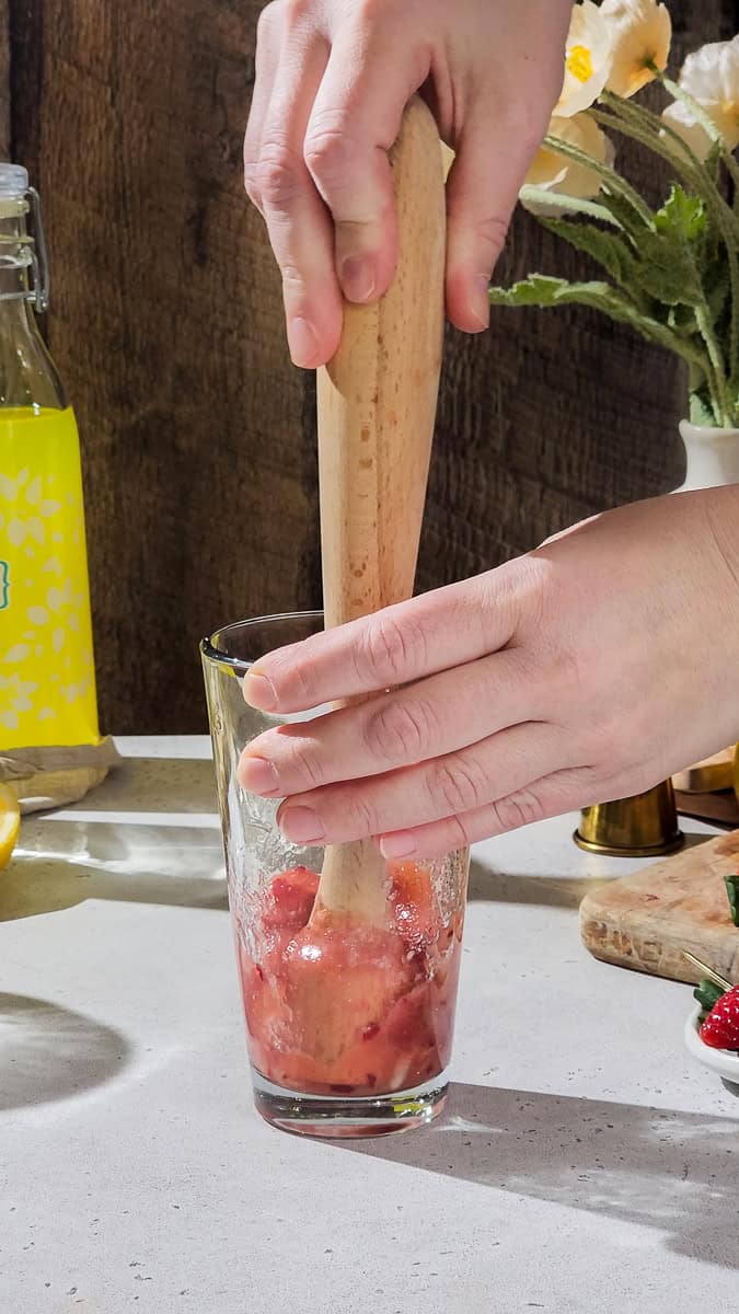 Hand using a muddler to mash up strawberries in a cocktail shaker.