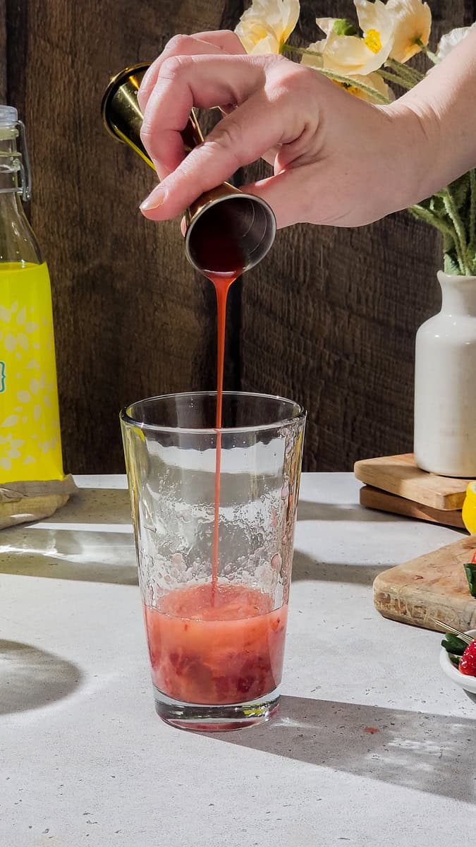 Hand adding strawberry syrup to a cocktail shaker.