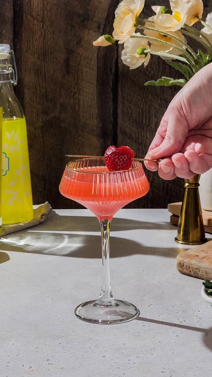 Hand adding a heart-shaped strawberry garnish to a red cocktail in a coupe glass.