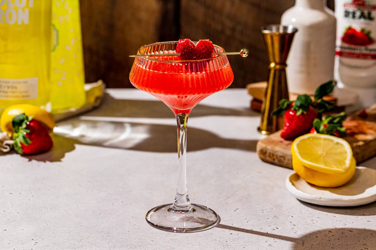 Side view of a Strawberry Lemon Martini in a coupe glass with a strawberry garnish. Fresh strawberries and lemons are in the background along with liquor bottles and a jigger.